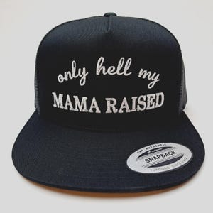 May include: Black trucker hat with the embroidered text "only hell my MAMA RAISED" in white script. The hat features a mesh back and a flat brim. A "Yupoong Snapback" sticker is on the brim.