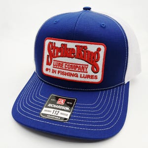 May include: A blue and white trucker hat with a curved brim. The hat features a rectangular patch with the words "Strike King Lure Company" in red and white. The text "#1 in Fishing Lures" is also visible. The hat is adjustable.