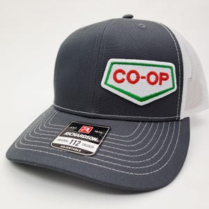May include: A charcoal gray trucker hat with a white mesh back and a curved brim. The hat features a patch with the text "CO-OP" in red letters on a white background, outlined in green. A black tag reads "RICHARDSON ORIGINAL 112 TRUCKER ADJUSTABLE".