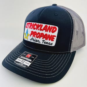 May include: A navy blue trucker hat with a gray mesh back. The front features a patch with the words "STRICKLAND PROPANE" in red, with "Arlen, Texas" below. The hat has a curved brim and a Richardson tag.
