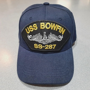 May include: Navy blue baseball cap featuring a patch with the text "USS BOWFIN" in gold lettering. The patch also includes the submarine's designation "SS-287" and a detailed emblem. The cap has a curved brim and a button on top.