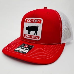 May include: Red and white trucker hat with a curved brim. The hat features a patch with the text "CO-OP" above a black pig illustration and the words "SWINE FEEDS" below. The hat is adjustable and has a white mesh back.