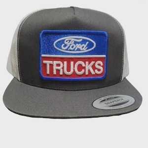 May include: A gray trucker hat with a flat brim and a white mesh back. The front features a rectangular patch with the Ford logo and the word "TRUCKS" in white lettering on a blue and red background.