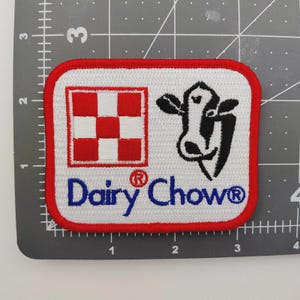 May include: Embroidered patch with a white background, red border, and the words "Dairy Chow" in blue. The patch features a red and white checkerboard design and a black and white cow graphic.