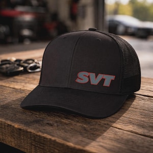 May include: Black baseball cap with a mesh back and the letters "SVT" in gray with a red outline. The cap is sitting on a wooden surface.