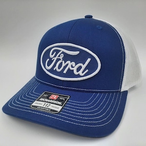 May include: A blue and white trucker hat with the Ford logo embroidered on the front. The hat features a white mesh back and a curved brim with white stitching. The Richardson 112 Trucker hat is adjustable.