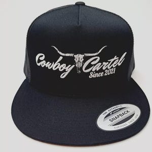 May include: Black trucker hat with a flat brim and mesh back. The hat features the text "Cowboy Cartel Since 2021" in white script, along with a steer skull graphic. A "Snapback" sticker is on the brim.