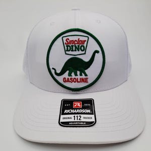 May include: White trucker hat with a green and white Sinclair Dino logo patch. The patch features a green dinosaur and the words "Sinclair DINO GASOLINE". The hat has a Richardson patch on the front.