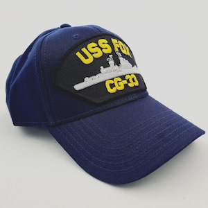 May include: Navy blue baseball cap featuring a patch with the text "USS FOX CG-33" in yellow lettering. The patch also displays a white illustration of a ship on a black background. The cap has a curved brim and a structured crown.
