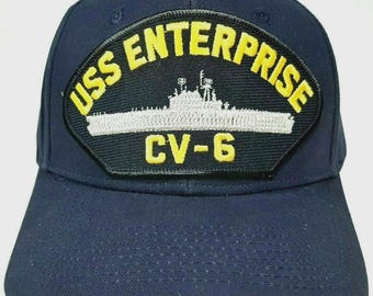 Pode incluir: Boné de beisebol azul marinho com um patch com o texto "USS ENTERPRISE CV-6" em amarelo e branco. O patch também inclui uma imagem de um navio. O boné é feito de um tecido resistente.