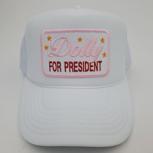 May include: White trucker hat with a curved brim and mesh back. The front features a rectangular patch with the words "Dolly FOR PRESIDENT" in pink and red, with gold star accents. A fun, novelty item.