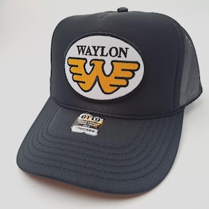May include: Black trucker hat with a mesh back and a curved brim. The front features an oval patch with the name "WAYLON" above a stylized "W" logo in yellow and white. The hat includes a "OTTO" tag.