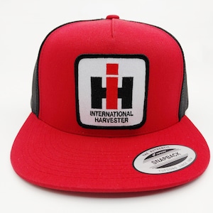 May include: Red and black snapback hat with the "International Harvester" logo on the front. The logo is a white square with a black and red emblem and the words "INTERNATIONAL HARVESTER" below it. The hat has a curved brim and a snap closure.