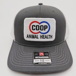 May include: Gray and white trucker hat with a curved brim. The hat features a white patch with the word "COOP" in red and blue circles, and the words "ANIMAL HEALTH" below. The hat has a Richardson tag.