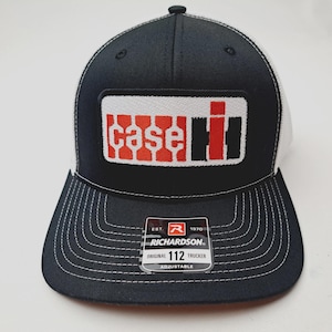 May include: Black and white trucker hat with a rectangular patch featuring the Case IH logo in red and black. The hat has a curved brim with white stitching and a white mesh back. The tag reads "Richardson 112 Trucker".