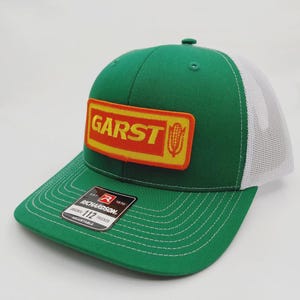 May include: Green and white trucker hat with the word "GARST" in orange letters on a yellow rectangle. The hat has a curved brim and a white mesh back. The Richardson brand tag is visible.