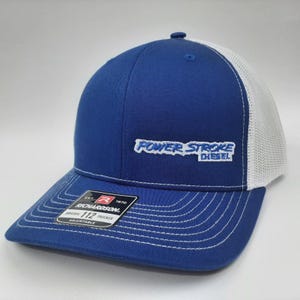 May include: A blue trucker hat with a white mesh back. The hat features the embroidered text "POWER STROKE DIESEL" in white and blue. The brim is curved with white stitching, and a Richardson tag is visible.