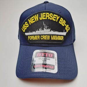 May include: Navy blue baseball cap with a patch featuring the text "USS NEW JERSEY BB-62 FORMER CREW MEMBER" and a ship illustration. The cap has a curved brim and a "OTTO" label.