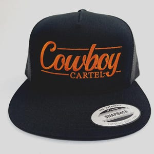 May include: Black trucker hat with the orange embroidered text "Cowboy Cartel". The hat features a flat brim, a mesh back, and a snapback closure. A small white tag with black text is on the front.