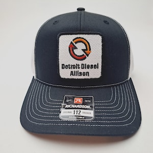 May include: Navy blue and white trucker hat with a square patch featuring the Detroit Diesel Allison logo. The hat has a curved brim, white mesh back, and adjustable closure. The Richardson brand tag is visible.