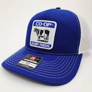 May include: A blue trucker hat with a white mesh back and a curved brim. The front features a patch with the text "CO-OP" above a black and white cow illustration and "DAIRY FEEDS" below. The hat is adjustable.