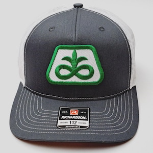 May include: A gray and white trucker hat with a green and white logo. The hat features a curved brim with white stitching and a white mesh back. The logo is a stylized design within a green border. A black and white tag reads "RICHARDSON ORIGINAL 112 TRUCKER ADJUSTABLE".