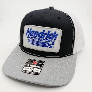 May include: A black, white, and gray trucker hat with the "Hendrick Motorsports" logo patch. The hat has a gray brim, a black front panel, and a white mesh back. The patch is rectangular with blue lettering and a checkered flag design.