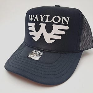 May include: Black trucker hat with the name "WAYLON" in white block letters above a stylized white "W" logo. The hat has a mesh back and a curved brim. A small tag on the brim reads "OTTO WORLD FAMOUS FOR FRONT ORM TRUCKER".