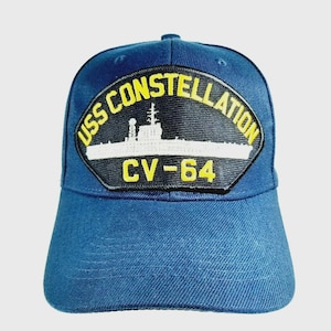 May include: A blue baseball cap featuring a patch with the text "USS CONSTELLATION" and "CV-64" in yellow and white. The patch depicts a white silhouette of a ship against a black background.