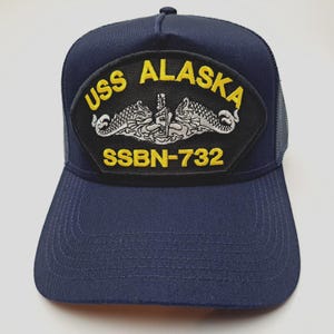 May include: Navy blue baseball cap with a mesh back. The front features a patch with the text "USS ALASKA" in yellow, along with the designation "SSBN-732" and a detailed emblem.