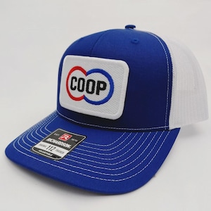 May include: A blue and white trucker hat with a white mesh back. The front features a rectangular patch with the word "COOP" in a red and blue design. The hat has a curved brim and a Richardson tag.