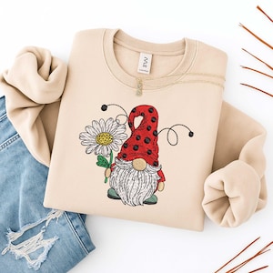May include: Beige crewneck sweatshirt featuring an embroidered gnome holding a daisy. The gnome has a red hat with black polka dots, a white beard, and green shoes. A gold necklace is draped on the sweatshirt. The sweatshirt is paired with blue jeans.