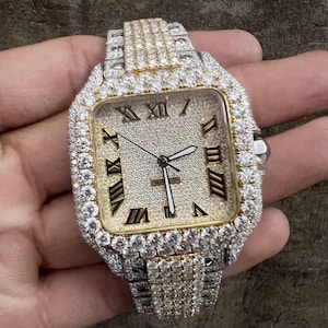May include: A luxury wristwatch with a square face and a silver and gold band. The watch face features black Roman numerals and silver hands. The watch is encrusted with numerous small, clear gemstones, creating a sparkling effect.