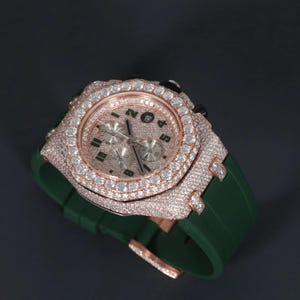 May include: A luxury wristwatch with a rose gold case and bezel, encrusted with numerous diamonds. The watch face is silver with green numerals. It has a green rubber strap.
