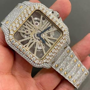 May include: A luxury wristwatch with a square face, adorned with many small diamonds. The watch features a gold bezel and a silver strap, also covered in diamonds. The watch hands are blue, and the inner mechanism is visible.