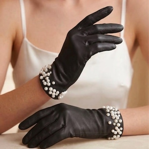 May include: A pair of black leather gloves featuring a cluster of white spherical pearls attached to the wrist area. The gloves are smooth in texture and extend past the wrist, with the pearl embellishments concentrated around the cuff opening.