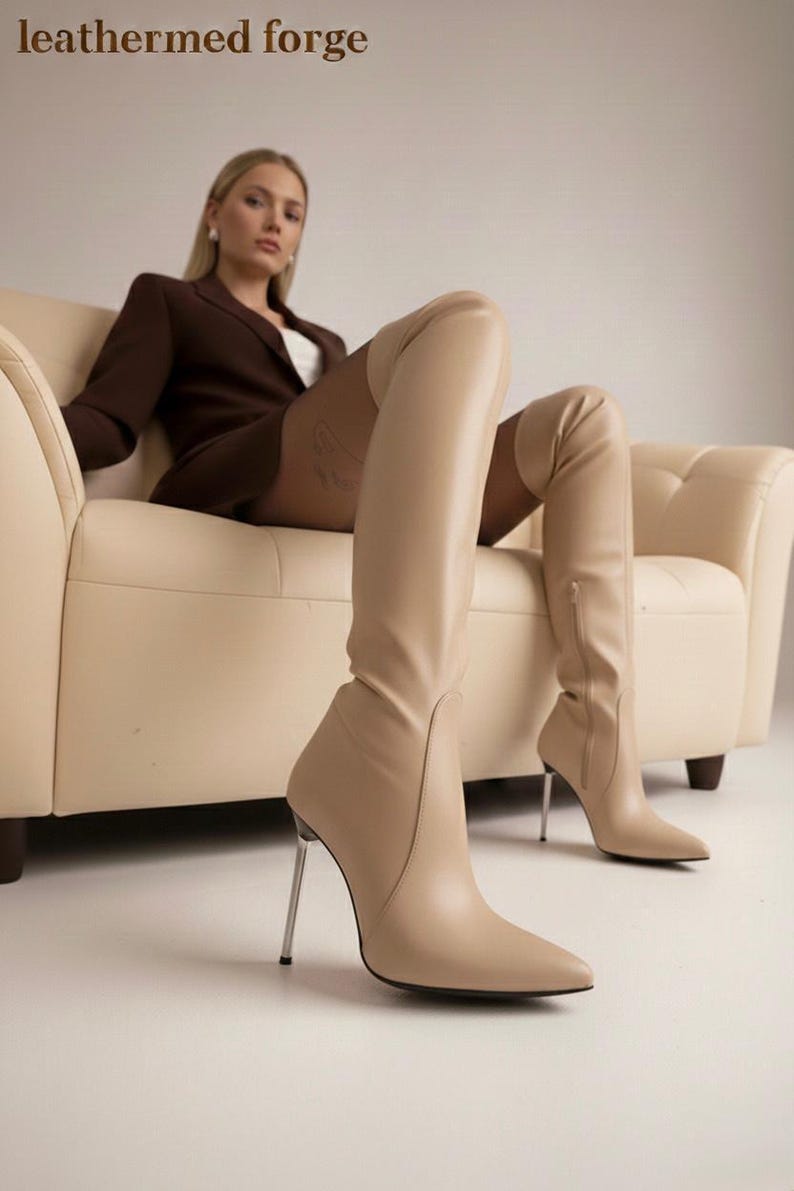May include: Beige leather thigh-high boots with pointed toes and stiletto heels. The boots have a zipper on the side and are worn by a person sitting on a cream-colored sofa. The text "leathered forge" is at the top of the image.