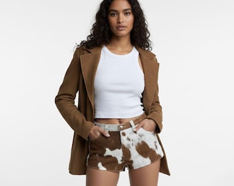 May include: A woman wearing a brown blazer over a white tank top and cowhide print shorts. The shorts have a brown and white pattern. The blazer is a solid brown color.
