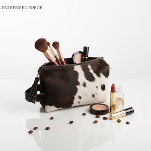 Puede incluir: Un neceser de maquillaje de piel de vaca con una borla negra, lleno de brochas de maquillaje y cosméticos. La bolsa es marrón y blanca, con cierre de cremallera. El texto "LEATHERMED FORGE" está en la esquina superior izquierda.