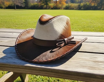 May include: A brown and white cowhide cowboy hat with a leather band and bow detail. The hat is sitting on a wooden bench outdoors, with a green grassy field in the background.