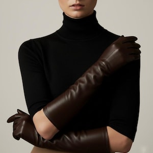 Peut inclure: Longs gants en cuir marron foncé. Les gants s'étendent au-delà des coudes. Le modèle porte un pull à col roulé noir. Les gants sont d'une couleur riche et unie et semblent être en cuir de haute qualité.