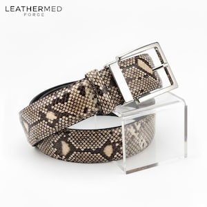 May include: A snakeskin-effect belt with a silver rectangular buckle. The belt is displayed on a clear acrylic stand. The brand name "LEATHERMED FORGE" is visible in the upper left corner.