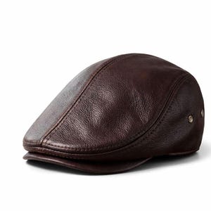 Peut inclure: Casquette plate en cuir marron foncé. Ce chapeau présente une couronne arrondie et une petite visière rigide. La surface est texturée et ornée de deux petits rivets métalliques. Un accessoire classique et intemporel, adapté à diverses occasions.