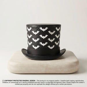 Puede incluir: Un sombrero de copa negro con un patrón repetitivo de murciélagos blancos en la corona cilíndrica. El sombrero tiene una banda negra lisa que rodea la base y un ala curva. El texto en la parte inferior dice: COPYRIGHT PROTECTED ORIGINAL DESIGN.