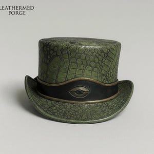 May include: A green top hat with a textured, crocodile-like pattern. The hat features a black band with a gold-colored eye-shaped design. The text "LEATHERMED FORGE" is visible in the upper left corner. The hat has a curved brim.