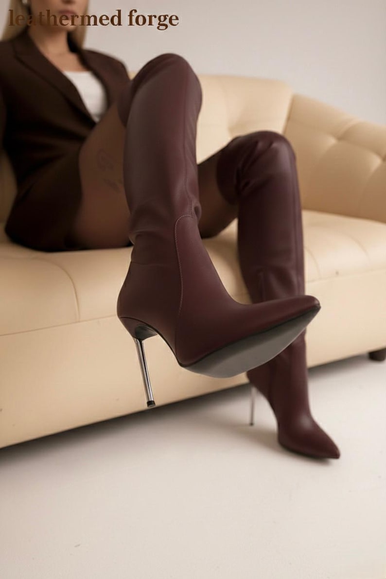 May include: Burgundy leather knee-high boots with stiletto heels. The boots have a pointed toe and a metallic silver heel. The image also shows the text "leathermed forge".