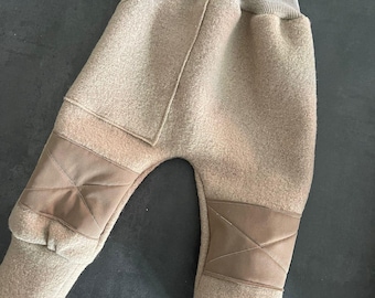 Puede incluir: Pantalones de color beige con una cintura alta acanalada y un tejido texturizado similar al fieltro. La prenda incluye rodilleras reforzadas de textura lisa con un patrón de cruz cosido y puños acanalados en los tobillos para un ajuste estructurado.