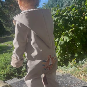Può includere: Una tuta intera con cappuccio color beige, maniche lunghe e polsini a coste su polsi e caviglie. Il retro del capo presenta una stampa grafica colorata raffigurante un muso di leone con una criniera stilizzata nei toni dell'arancione e del blu.