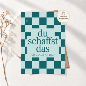 Könnte beinhalten: Eine rechteckige Grußkarte mit einem blaugrünen und hellblauen Schachbrettmuster. Der mittige Text lautet "du schaffst das" und "ICH GLAUB AN DICH" in Dunkelblaugrün. Ein kleines Herzsymbol ist oben links und ein runder Aufkleber zeigt "AUF WUNSCH MIT UMSCHLAG".