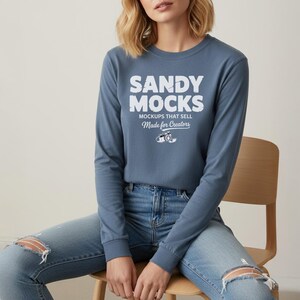 May include: A long-sleeved, dusty blue shirt with the text "SANDY MOCKS" in bold white letters. Below the text are the words "MOCKUPS THAT SELL" and "Made for Creators". The shirt is paired with ripped blue jeans.
